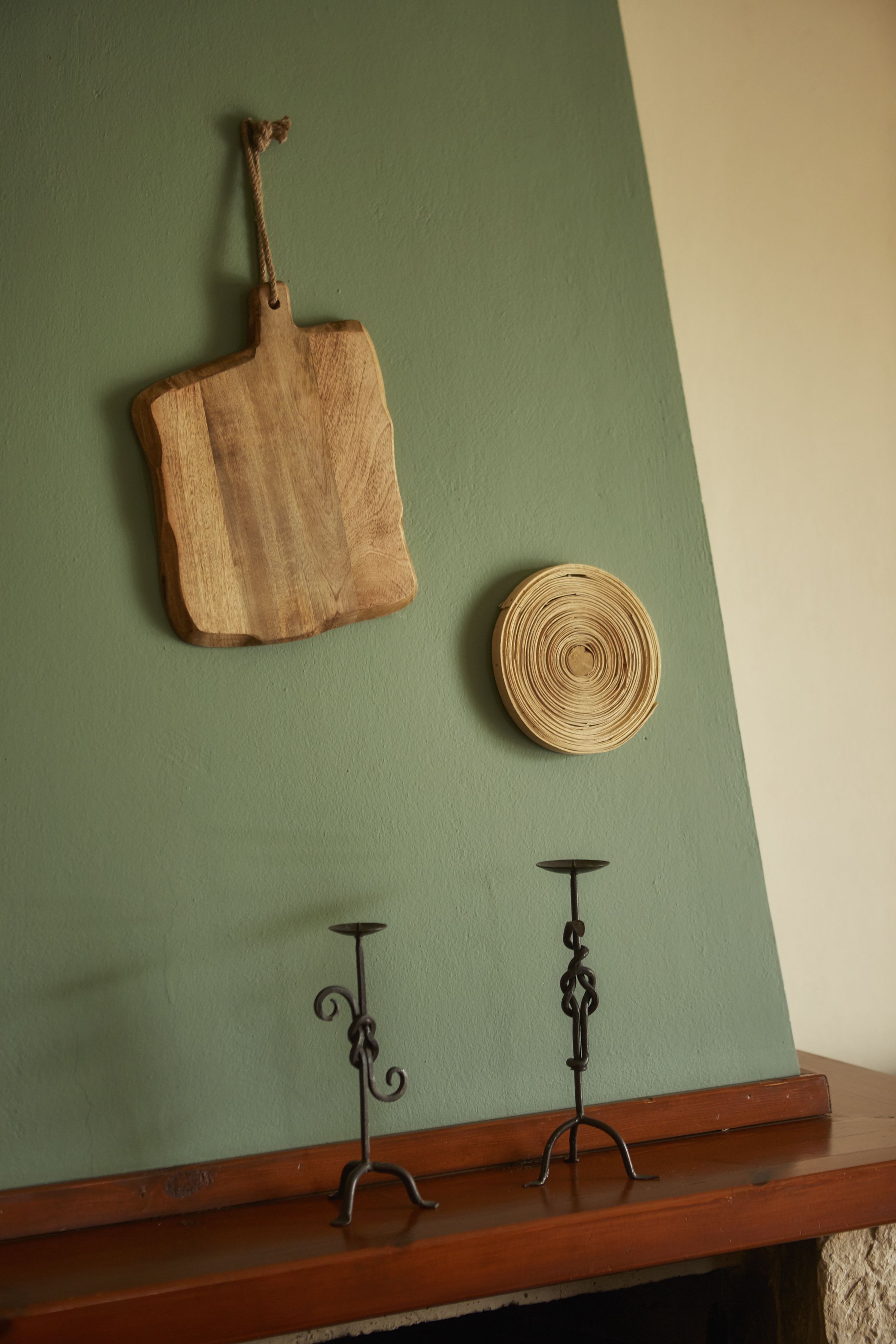 Une planche à découper en bois accrochée à un mur vert, une nappe ronde tissée placée en dessous, et deux bougeoirs posés sur une étagère en bois.
