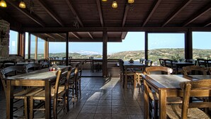 A restaurant with wooden tables and chairs, large windows offering a view of the hills, and hanging pendant lights. - Earino (Heraklion)