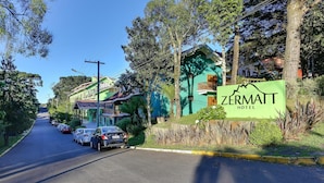 A hotel named Zermatt Hotel with a green facade, parked cars along the street, and lush greenery. - Zermatt Hotel (Gramado)