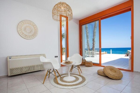 A modern room with a view of the beach, featuring a white radiator, two white chairs, a round table, and a woven floor mat.