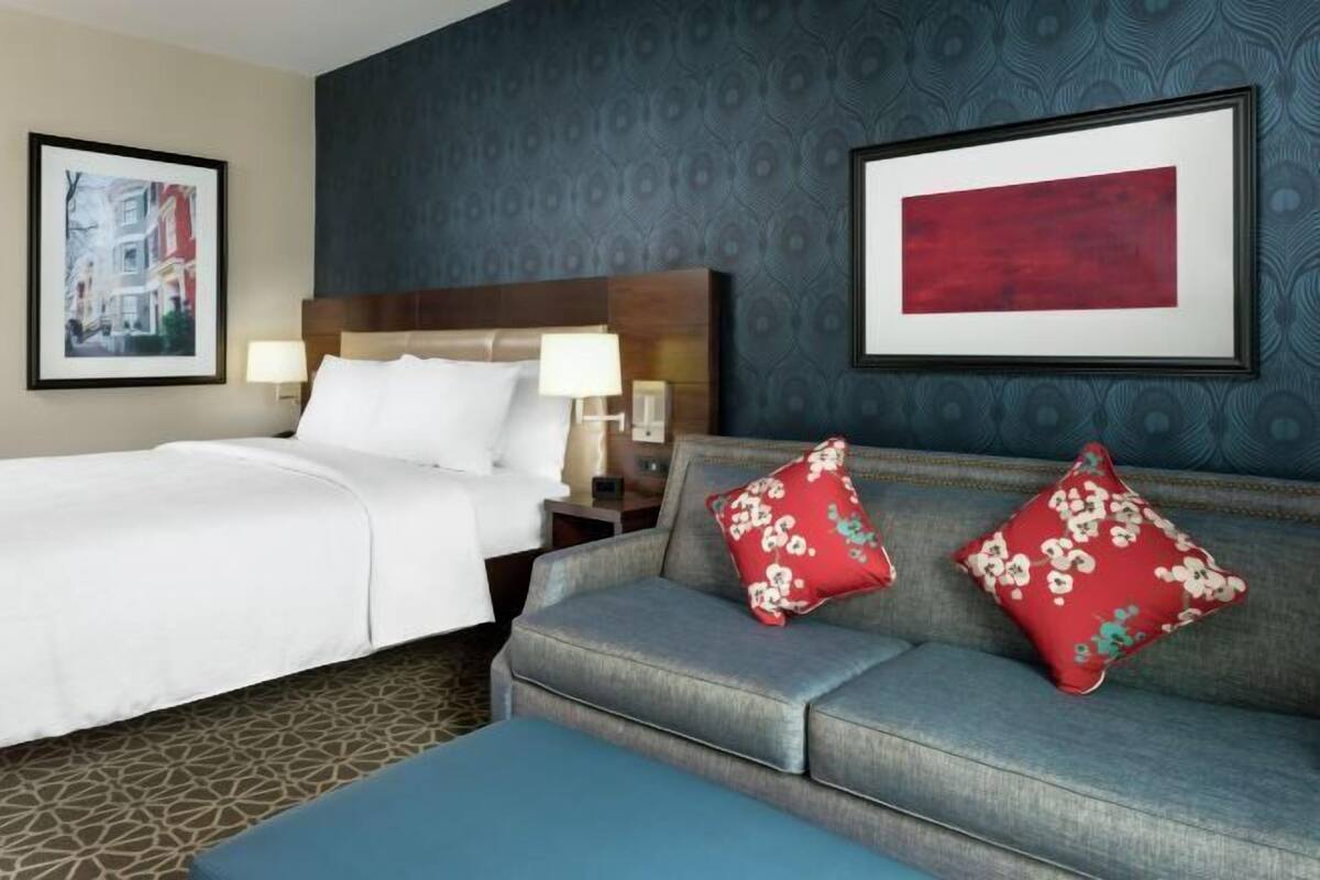 A hotel room with a bed, a sofa, two red floral pillows, a framed abstract painting, and a patterned wall.
