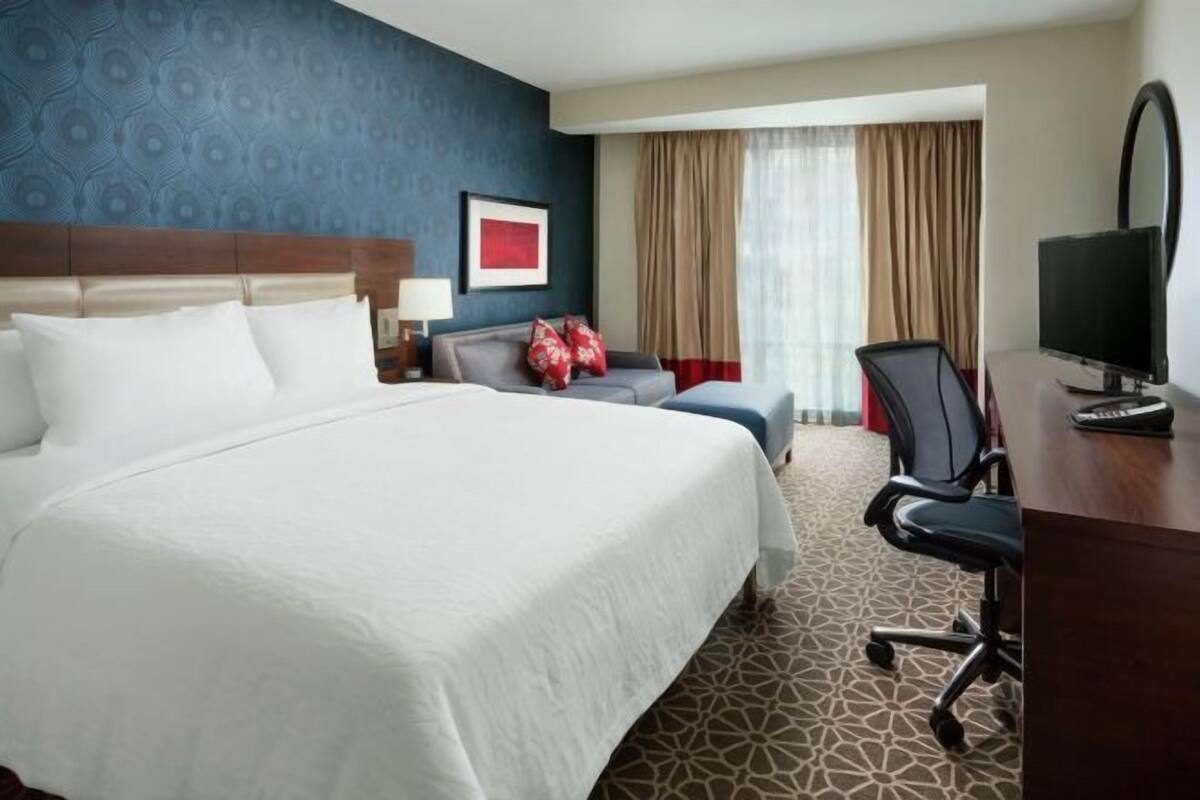A hotel room with a large bed, a desk with a computer, a chair, a TV, and a window with curtains.