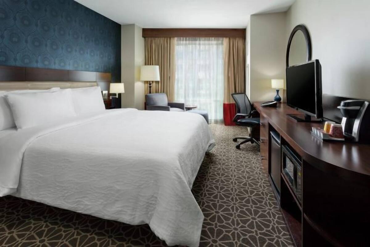 A hotel room with a large bed, a desk with a television, a chair, and a small table with a lamp.