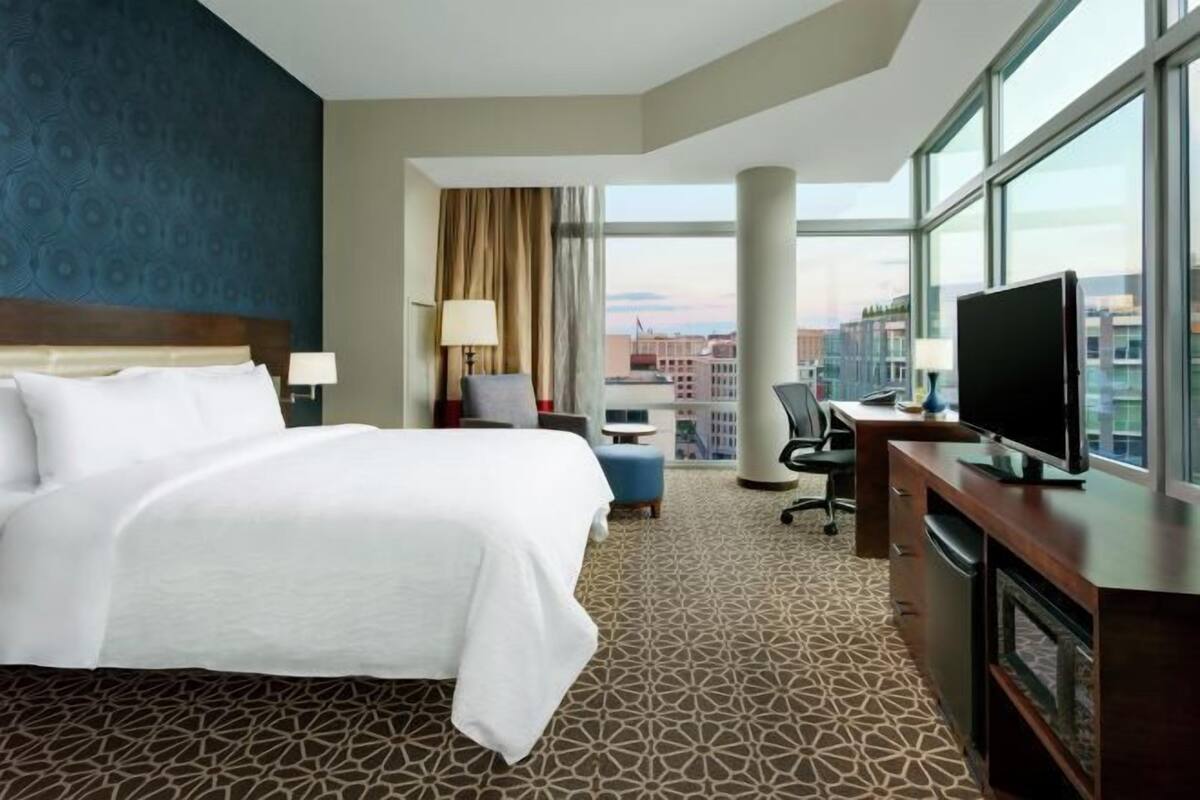 A hotel room with a large bed, a desk with a TV, a chair, and a view of the city through large windows.