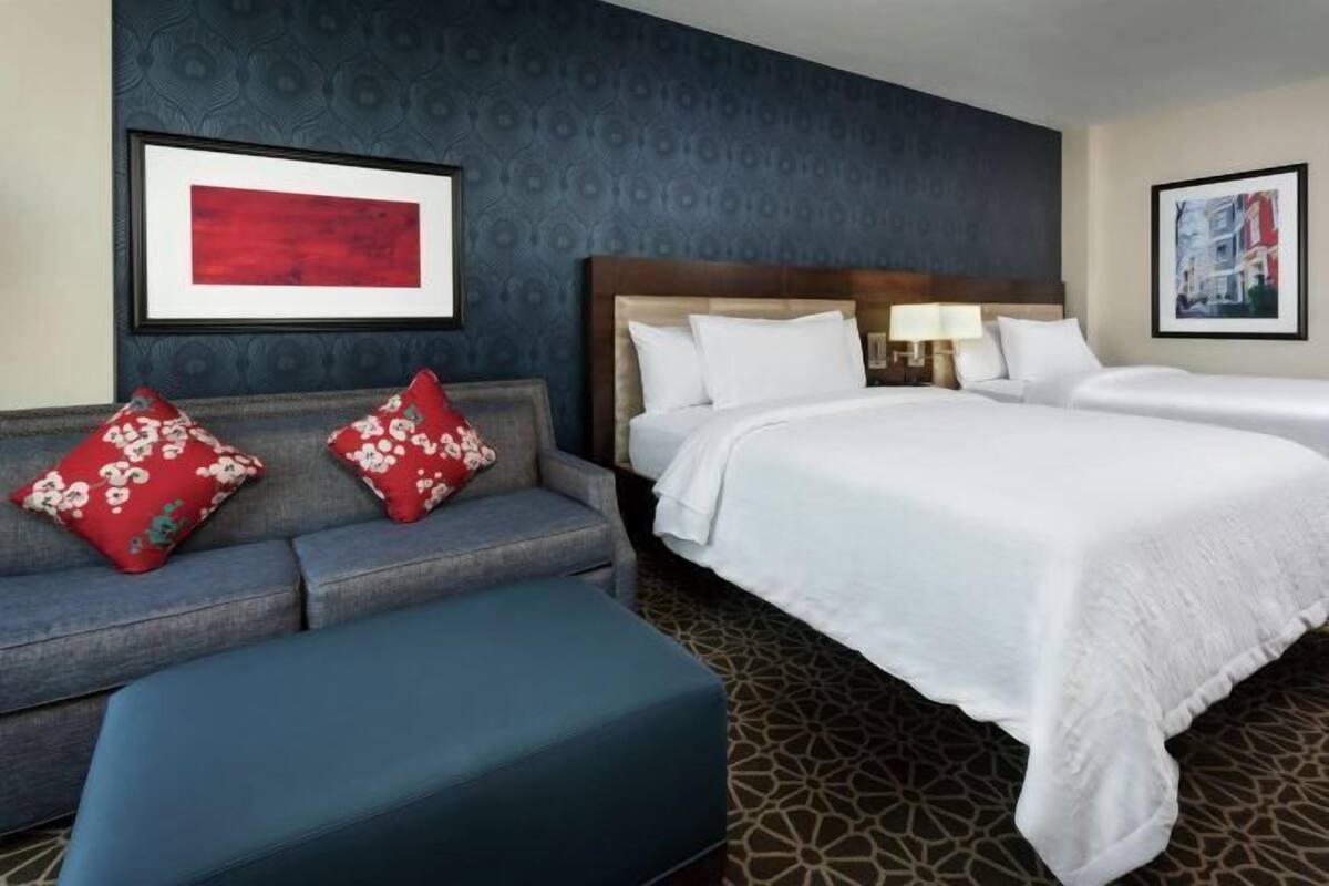 A hotel room with a sofa, two beds, a patterned wall, and framed artwork.