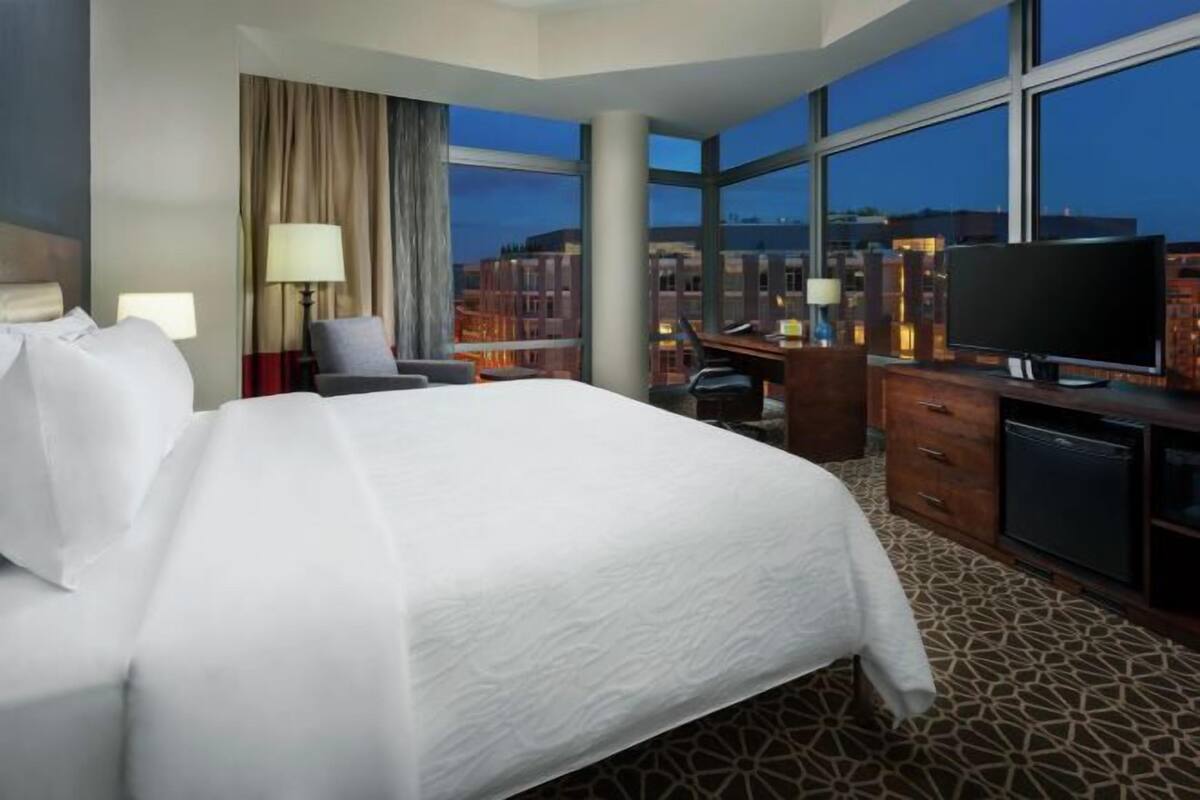 A hotel room with a large bed, a desk, a television, and a view of the cityscape through large windows.