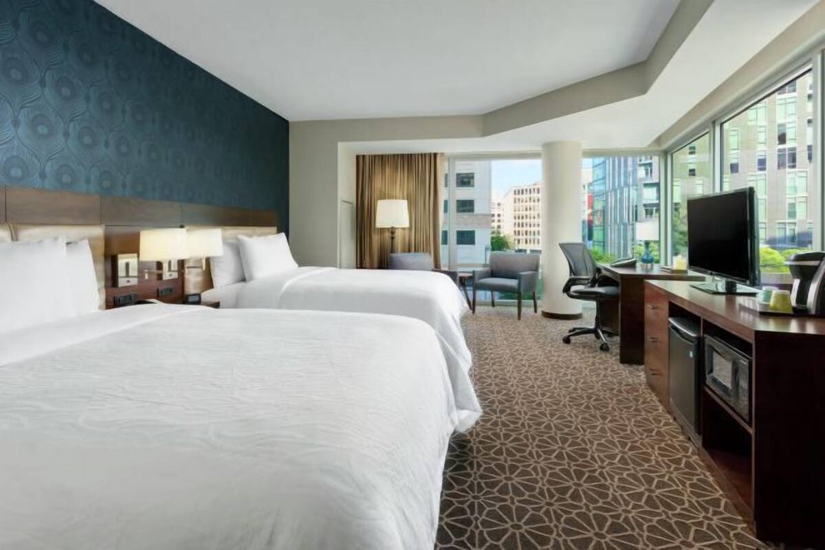 A hotel room with two beds, a desk with a TV, and a large window with a city view.