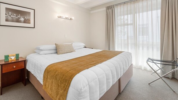 A hotel room with a large bed, a nightstand with a lamp, a framed picture on the wall, and a window with curtains. - The Parnell Hotel and Conference Centre (Auckland)