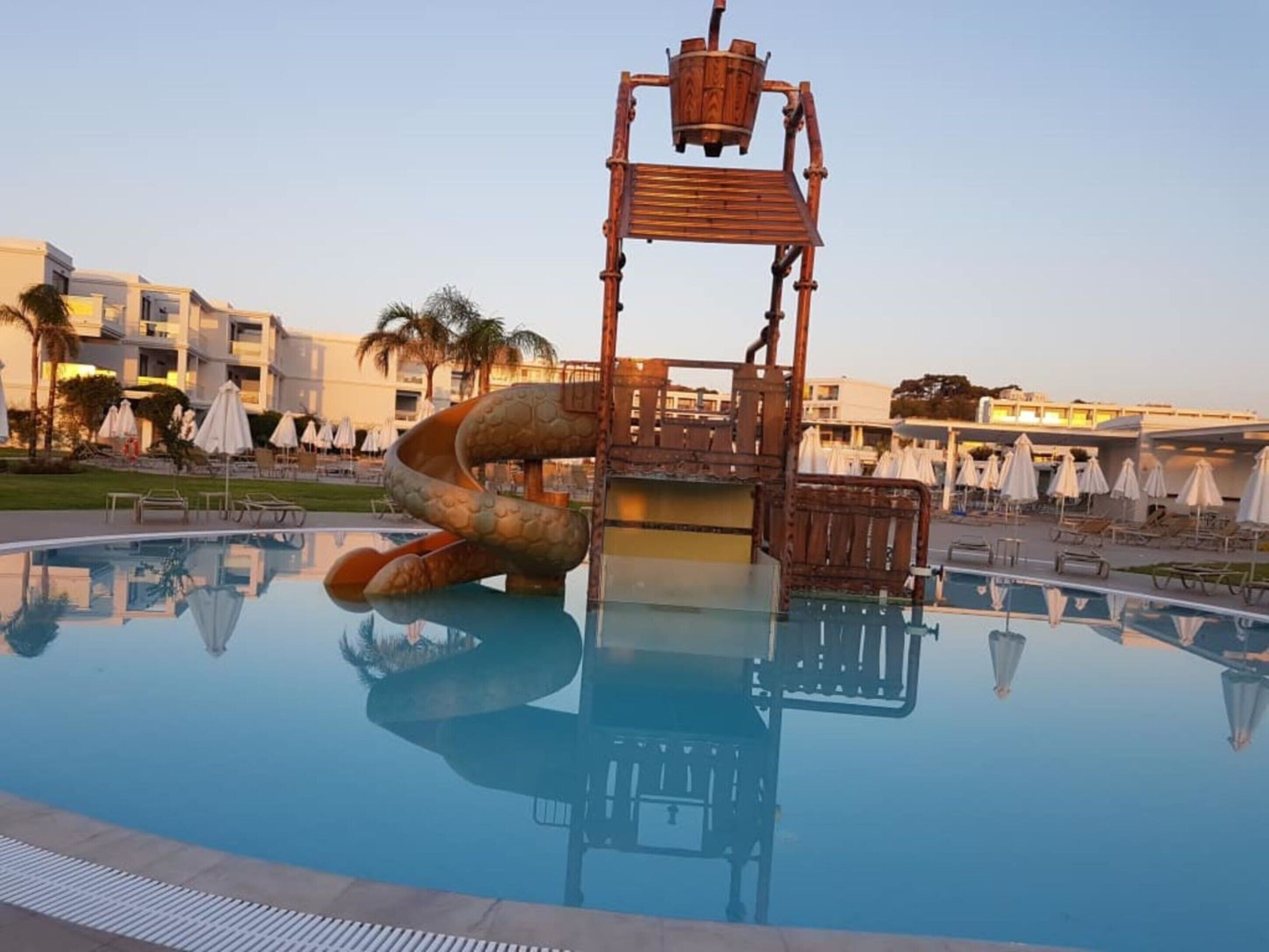 Piscina com estrutura de escorrega singular, rodeada por espreguiçadeiras e guarda-sóis, com edifícios de hotel ao fundo.