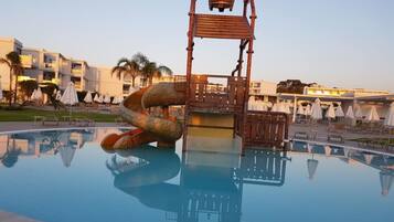 Piscina com estrutura de escorrega singular, rodeada por espreguiçadeiras e guarda-sóis, com edifícios de hotel ao fundo.