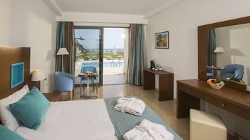 A hotel room with a bed, a desk, a chair, a small table, and a view of the pool and beach.