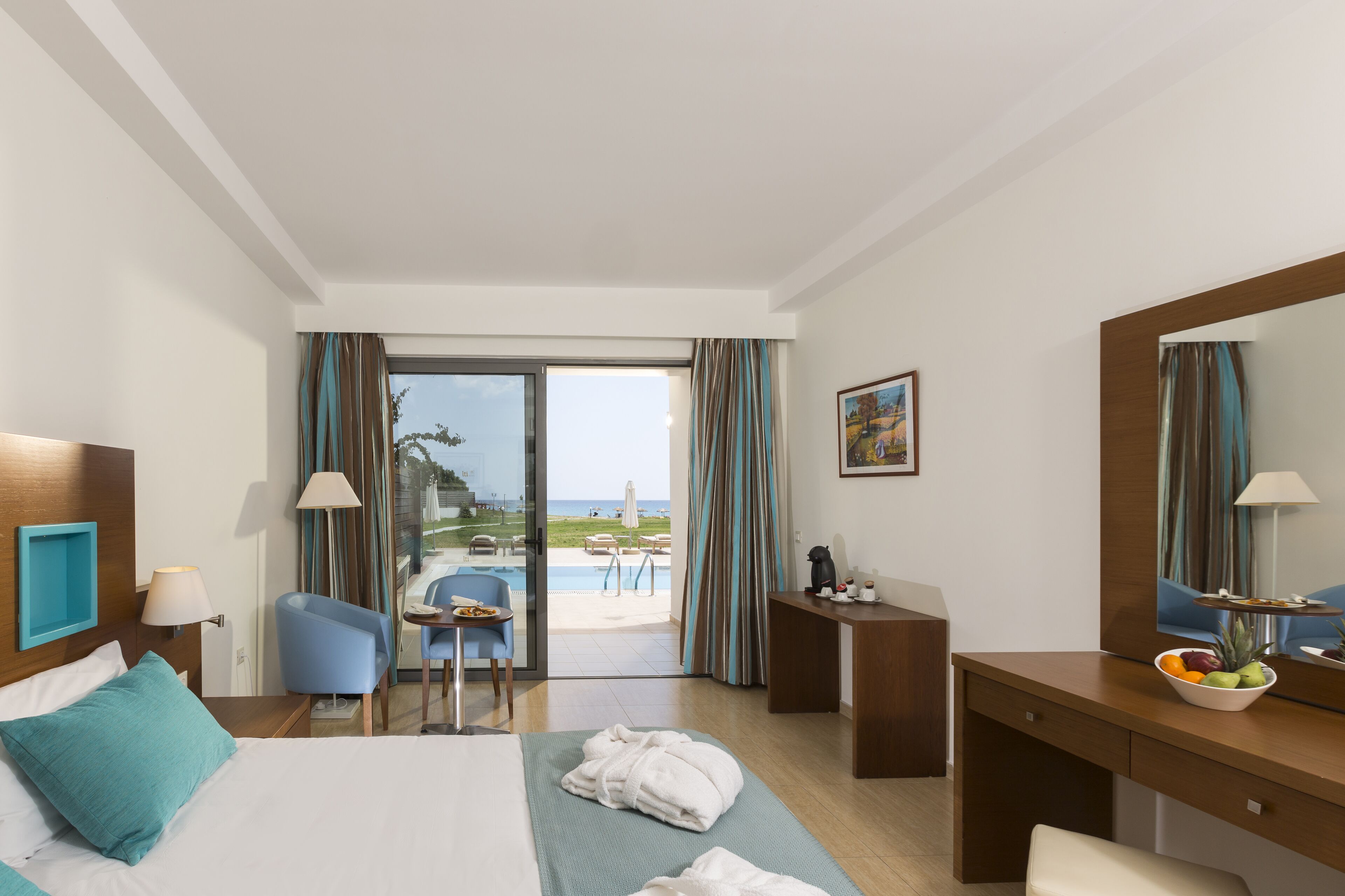 A hotel room with a bed, a desk, a chair, a small table with a bowl of fruit, and a view of the beach and pool through the glass door.