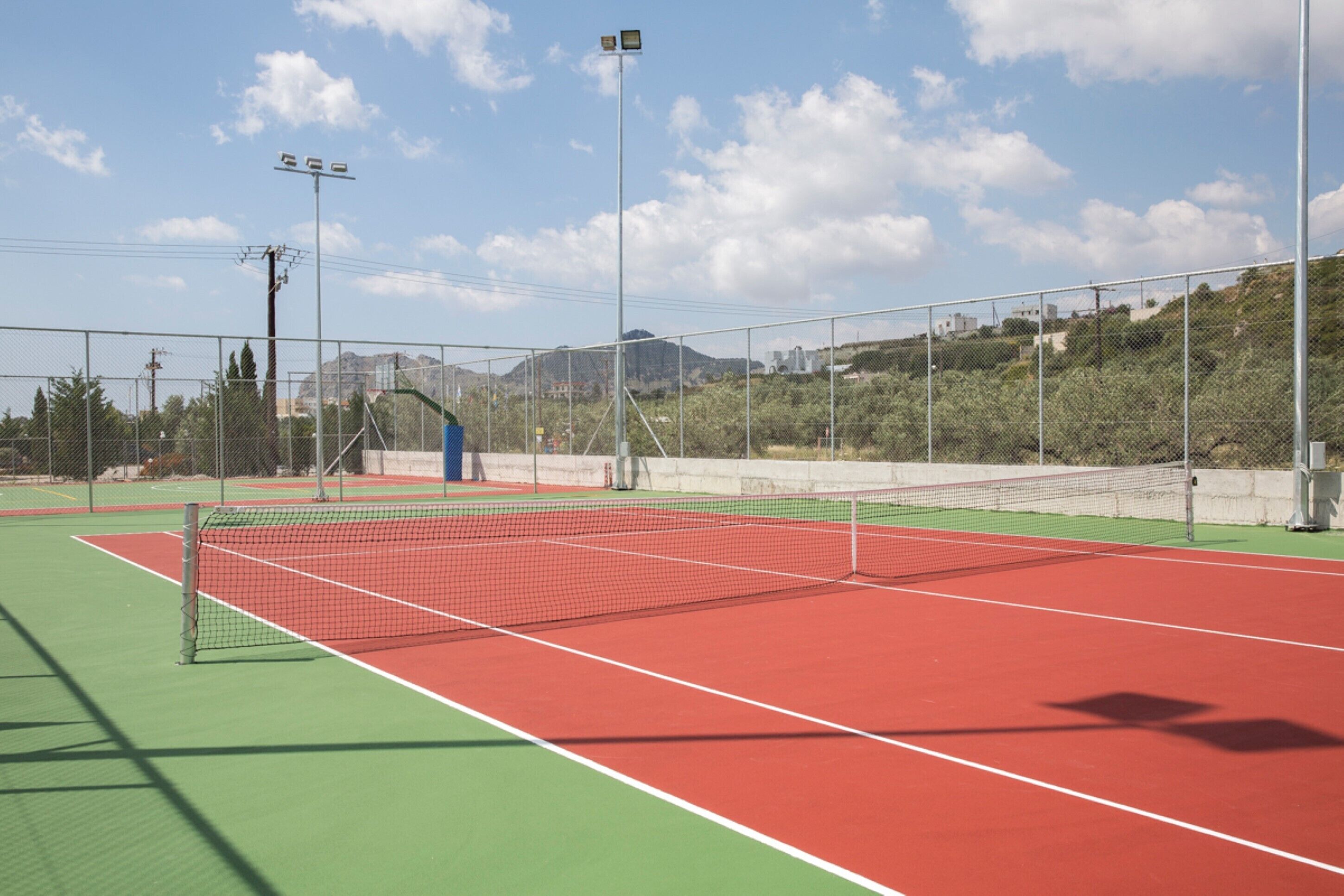 Una cancha de tenis con red, rodeada por una cerca, y un cielo despejado con nubes.