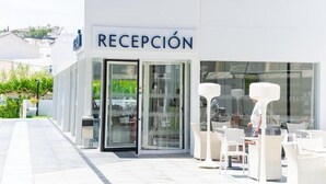 A hotel reception with a glass door entrance, outdoor seating area with white chairs and tables, and a clear view of the surrounding buildings.