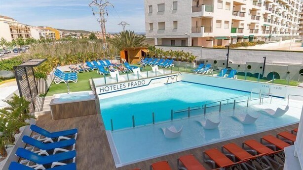 A hotel pool area with blue and orange lounge chairs, a sign reading "HOTELES PRADO," and a view of surrounding buildings.