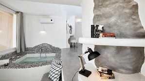 A modern living room with a stone wall, a small pool, a sofa, and a desk with books. - Suites of the Gods Cave Spa Hotel (Santorini)