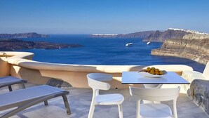 A terrace with white chairs and a table, overlooking a coastal landscape with cliffs and the sea. - Suites of the Gods Cave Spa Hotel (Santorini)
