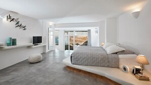 A modern bedroom with a bed, bedside lamp, books, and a TV on a shelf. - Suites of the Gods Cave Spa Hotel (Santorini)