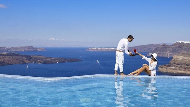 A couple by a pool with a view of the sea and cliffs. - Suites of the Gods Cave Spa Hotel (Santorini)