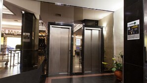 Vista interior de un vestíbulo de hotel con un mostrador de recepción, dos ascensores y una planta en maceta.