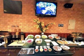 A buffet setup with various food items, a television mounted on the wall, and a brick wall background. - Fortune Hotel Deira (Dubai)