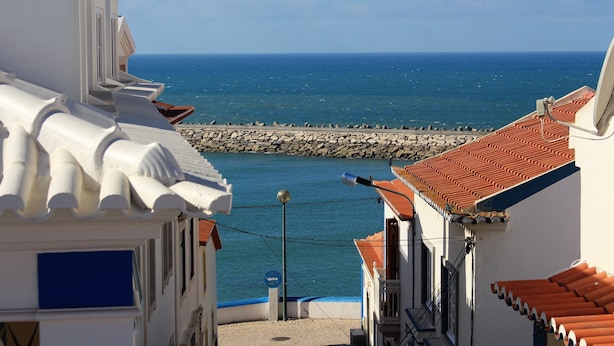 A coastal street with buildings, a rocky breakwater, and the sea in the background. - Villa Ana Margarida Residences (Mafra)