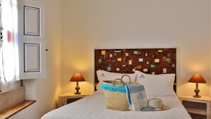 A neatly made bed with a patterned bedspread, a woven basket, and a headboard with a mosaic design. There are two bedside lamps and a small wooden nightstand. - Villa Ana Margarida Residences (Mafra)
