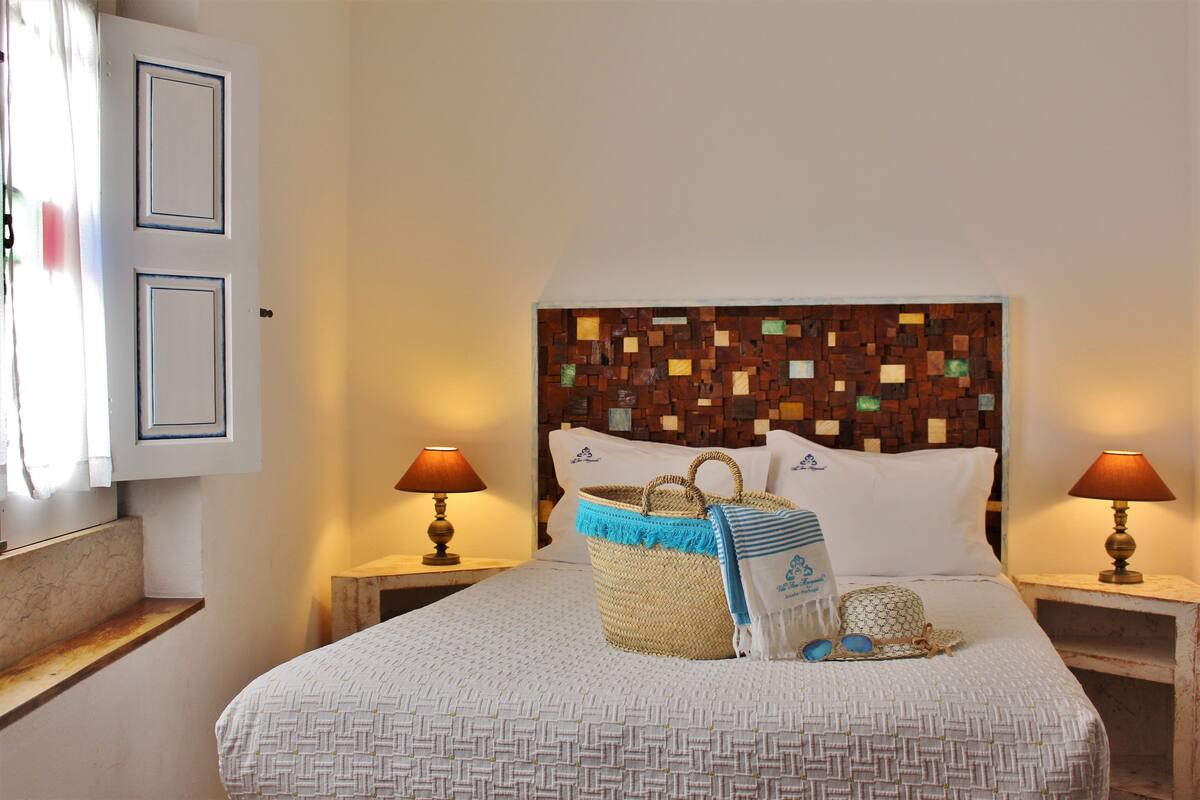 Een net opgemaakt bed met een bedsprei met patroon, een geweven mand en een hoofdbord met een mozaïek ontwerp. Er staan twee bedlampen en een klein houten nachtkastje.