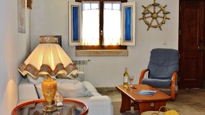 A living room with a sofa, a table with a lamp, a chair, a window with curtains, and a ship's wheel decoration. - Villa Ana Margarida Residences (Mafra)