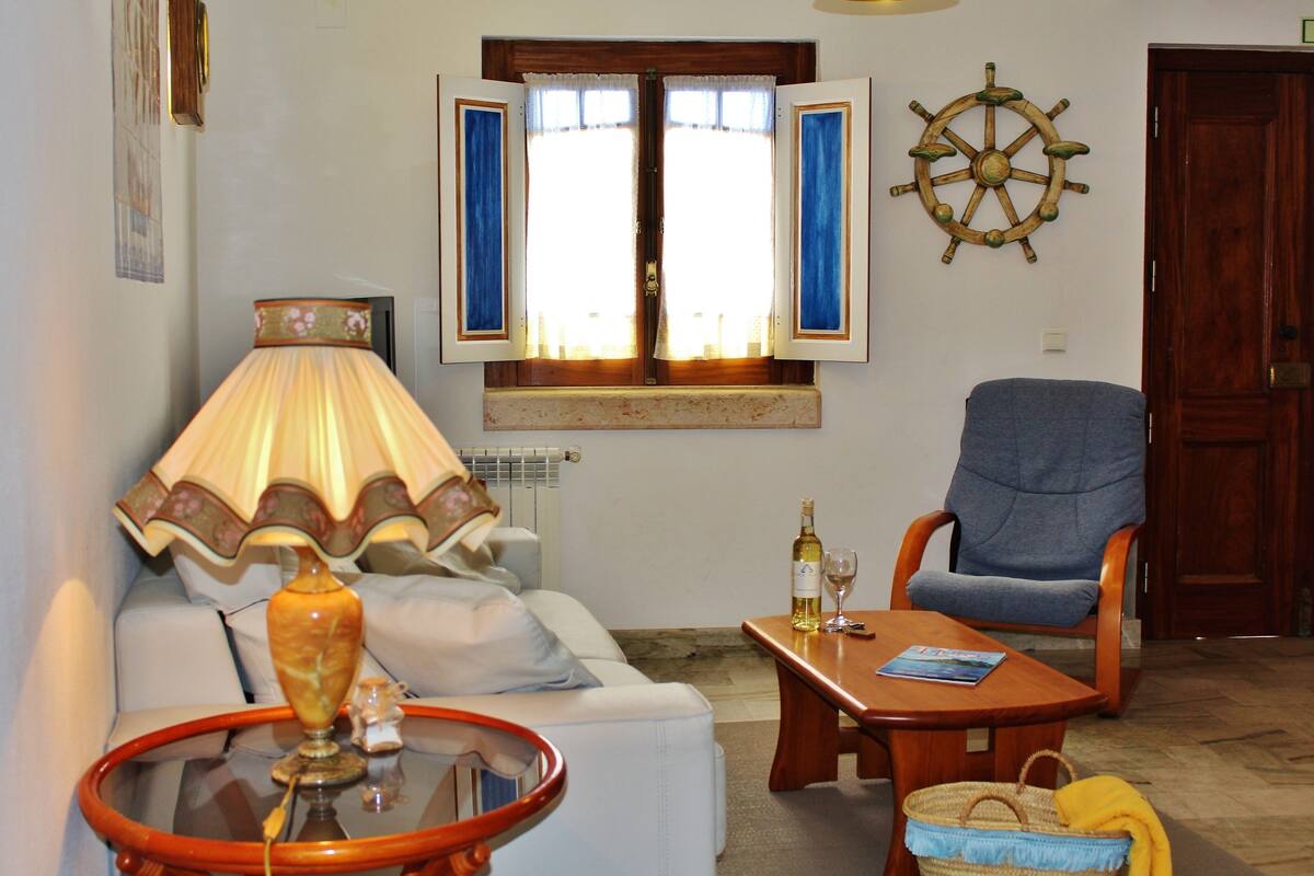 Een woonkamer met een bank, een tafel met een lamp, een stoel, een raam met gordijnen en een decoratie van een stuurrad.