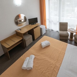 A hotel room with a bed, a desk with a computer, a chair, a small table, and a window with curtains.