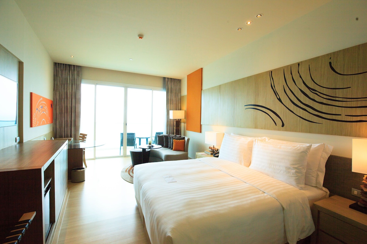 A modern hotel room with a large bed, a desk, and a seating area with a view of the outdoors.