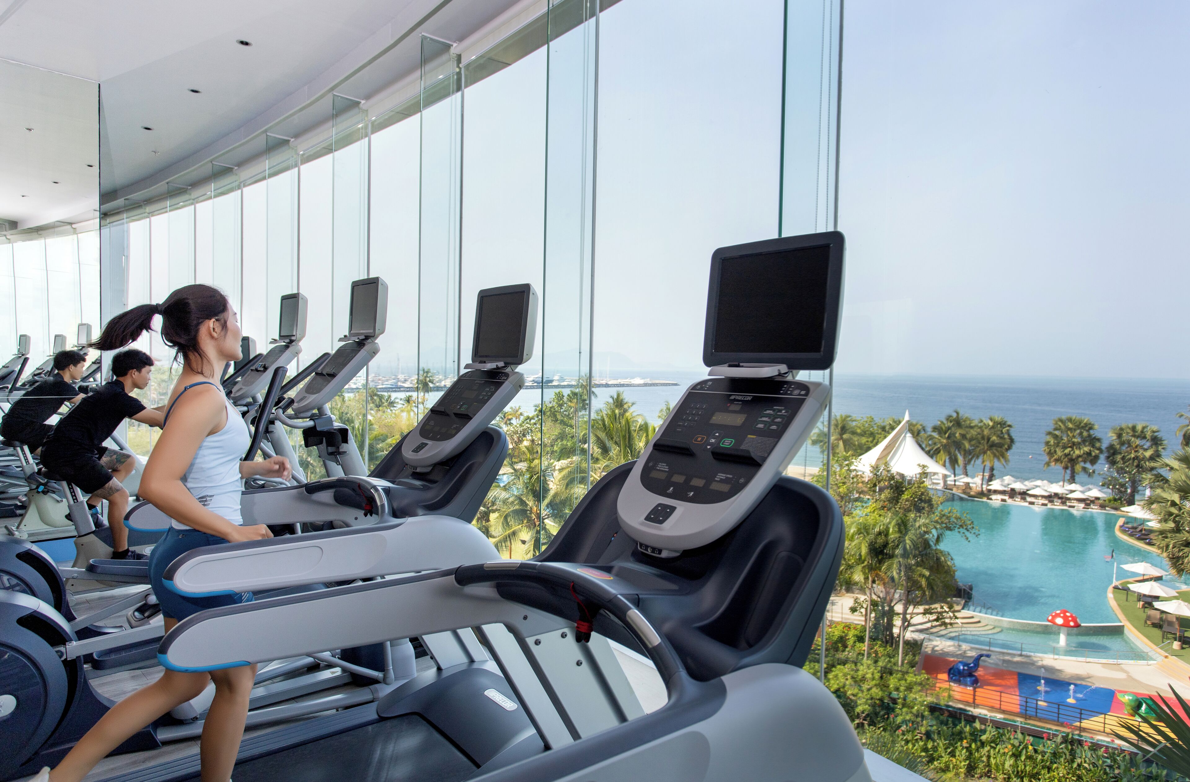 Une salle de sport équipée de tapis de course, avec vue sur une piscine et des palmiers.