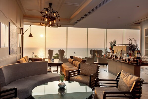 A modern lounge area with a curved sofa, armchairs, and a glass coffee table. A bar with a variety of bottles and glasses is visible in the background. - Mövenpick Siam Hotel Na Jomtien Pattaya (Sattahip)