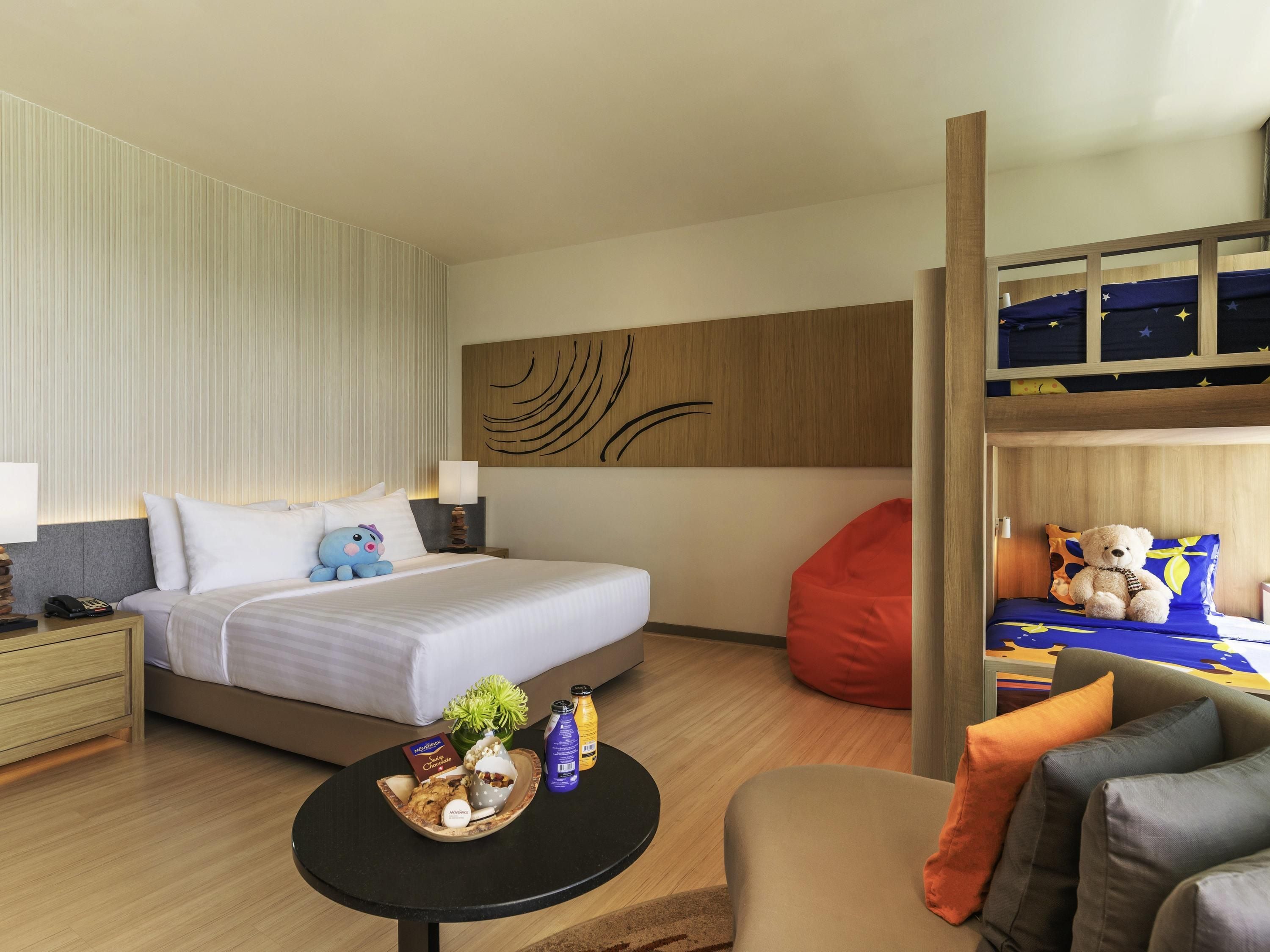 A hotel room with a bed, a nightstand, a bedside table, a round coffee table, a red bean bag, a sofa, and a bunk bed with a teddy bear.