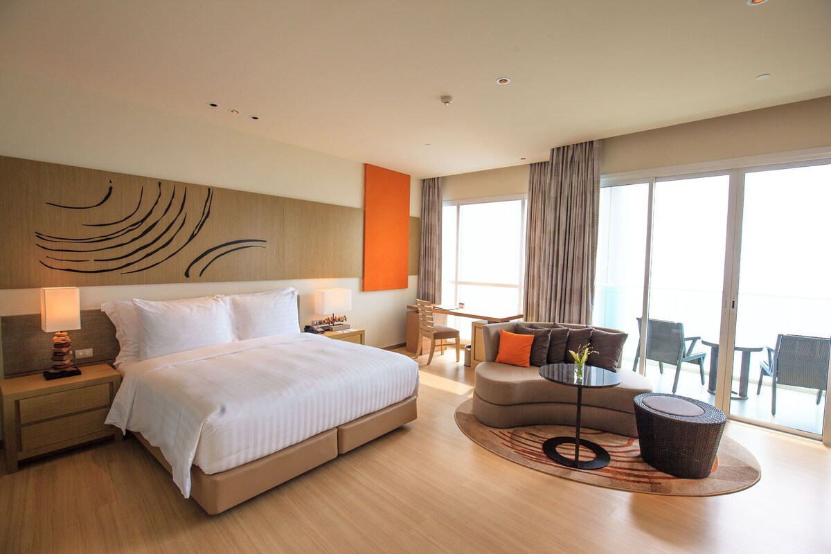 A modern hotel room with a large bed, a desk, and a seating area with a view of the outdoors.