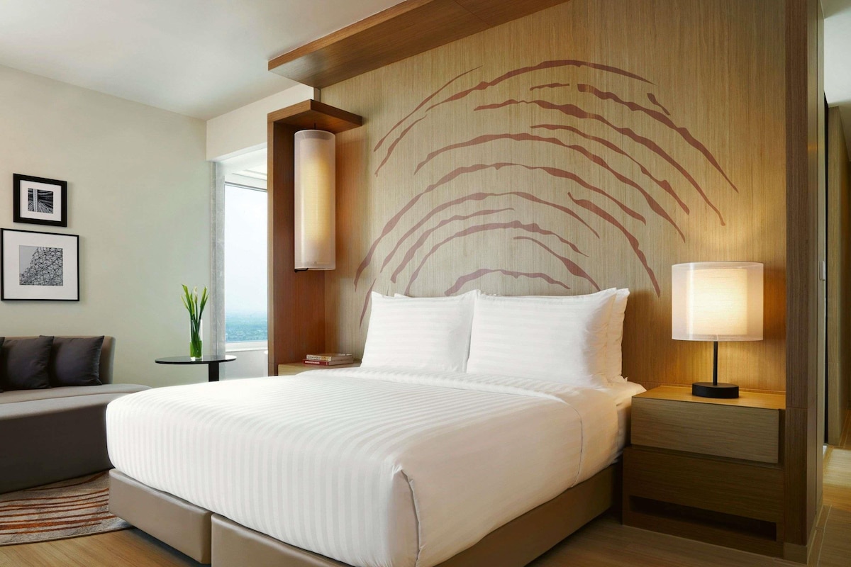 A modern hotel room with a large bed, a nightstand with a lamp, a sofa, and a wall with a unique design.