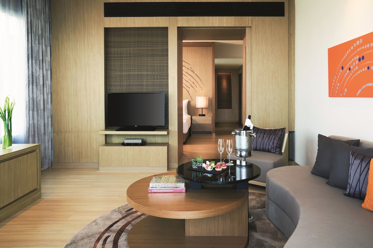 A modern hotel room with a flat-screen TV, a sofa, a coffee table with a book, and a dining area with a table set for two.