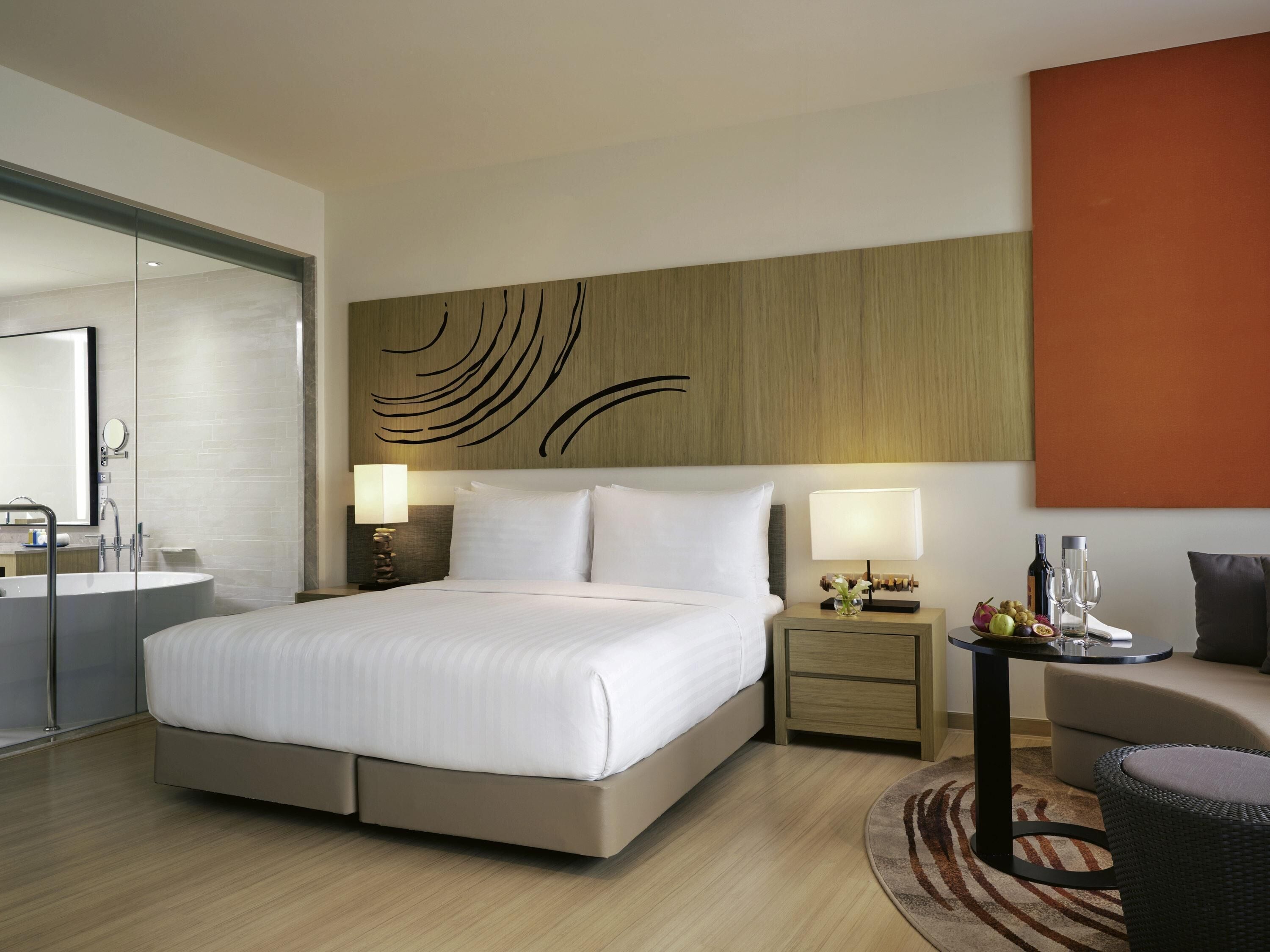 Habitación de hotel con cama, mesitas de noche, espejo, una mesa redonda con frutas y vasos, y una pared decorada con arte abstracto.