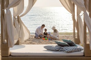 A beach setting with a family playing on the sand, a beach chair with pillows, and a view of the sea. - Mövenpick Siam Hotel Na Jomtien Pattaya (Sattahip)