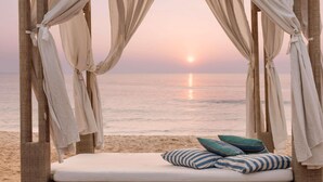 A wicker swing bed with white cushions and blue-striped pillows, overlooking a beach with a sunset. - Mövenpick Siam Hotel Na Jomtien Pattaya (Sattahip)