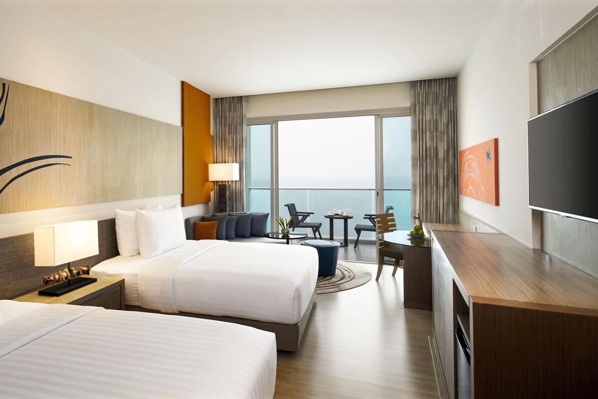 A hotel room with two beds, a desk, a chair, a small table, and a TV.