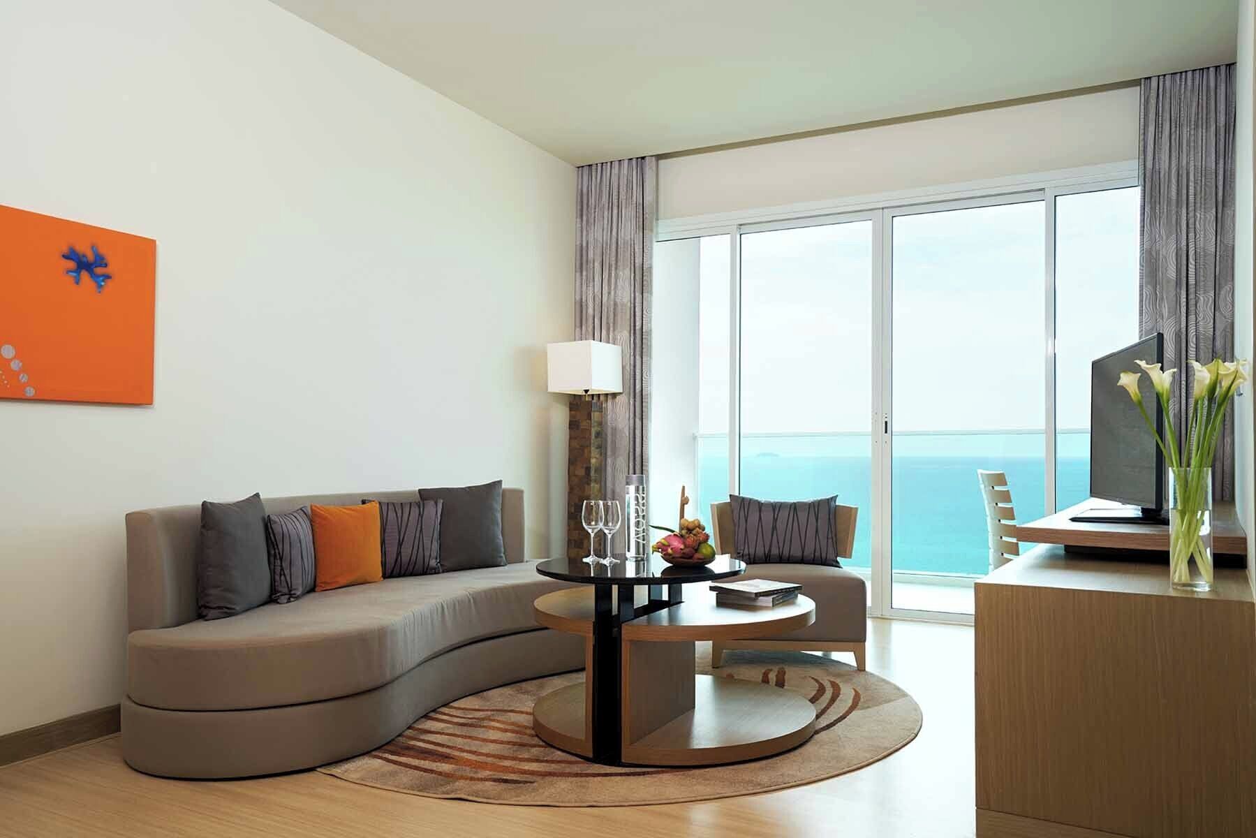 A modern living room with a curved sofa, a round coffee table, a TV on a wooden stand, and a view of the sea through large windows.