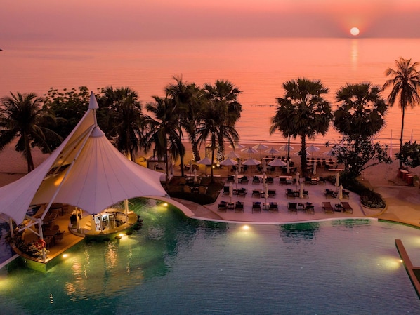 A resort pool with lounge chairs and umbrellas, palm trees, and a sunset view.