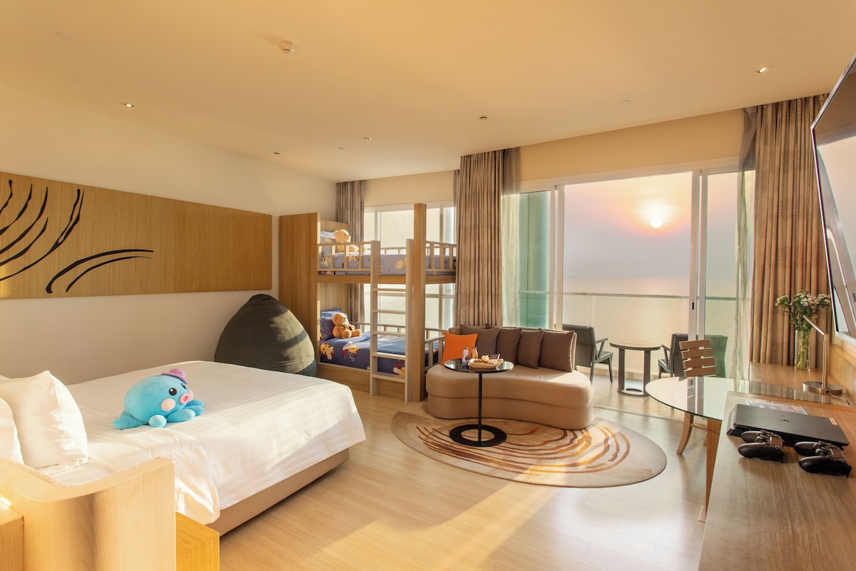 A hotel room with a bed, a sofa, a dining table, and a balcony with a view of the sunset.