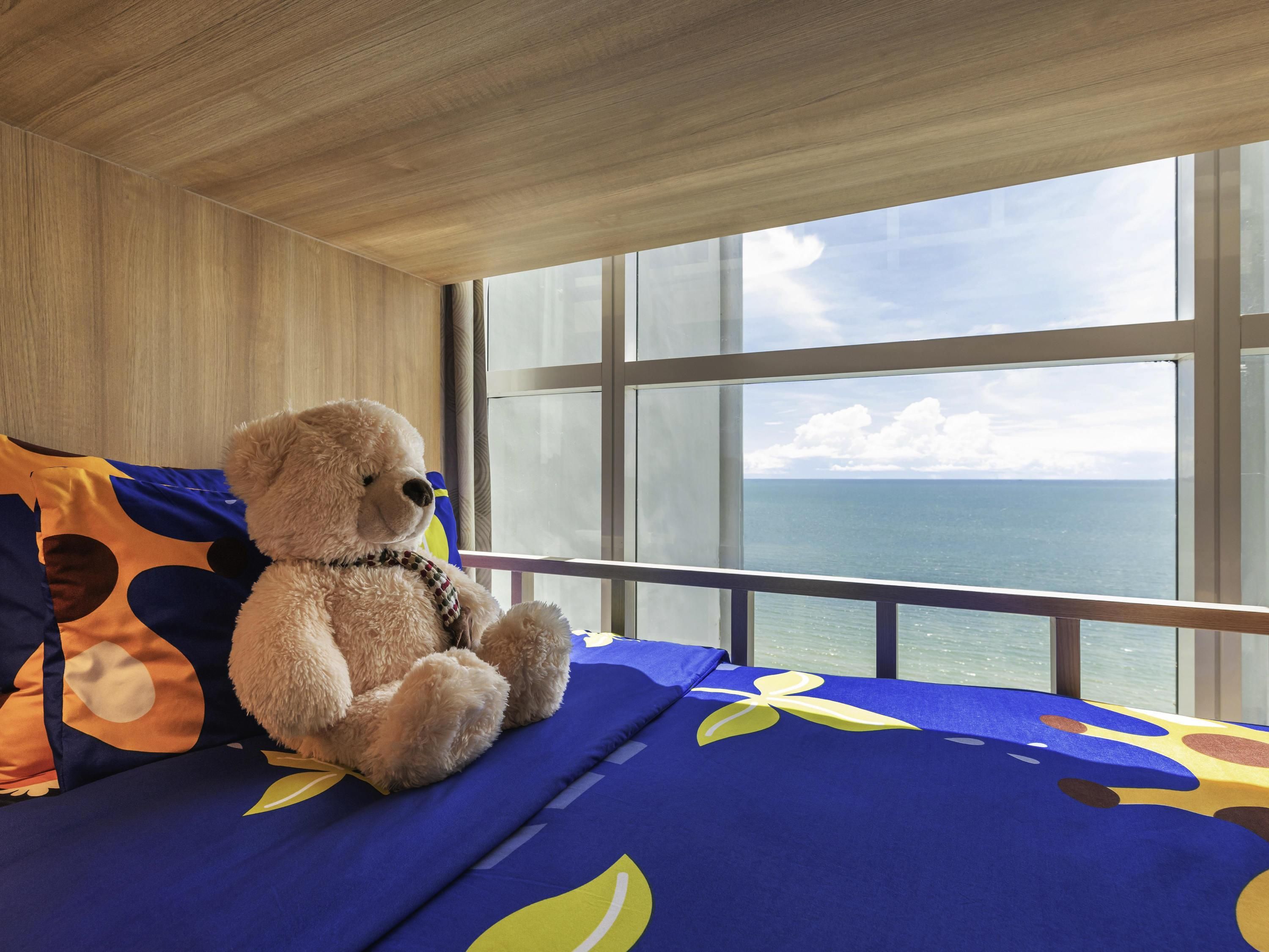 Een kamer met een groot raam, een bed met een deken met blauw en geel dessin, en een teddybeer op het bed.