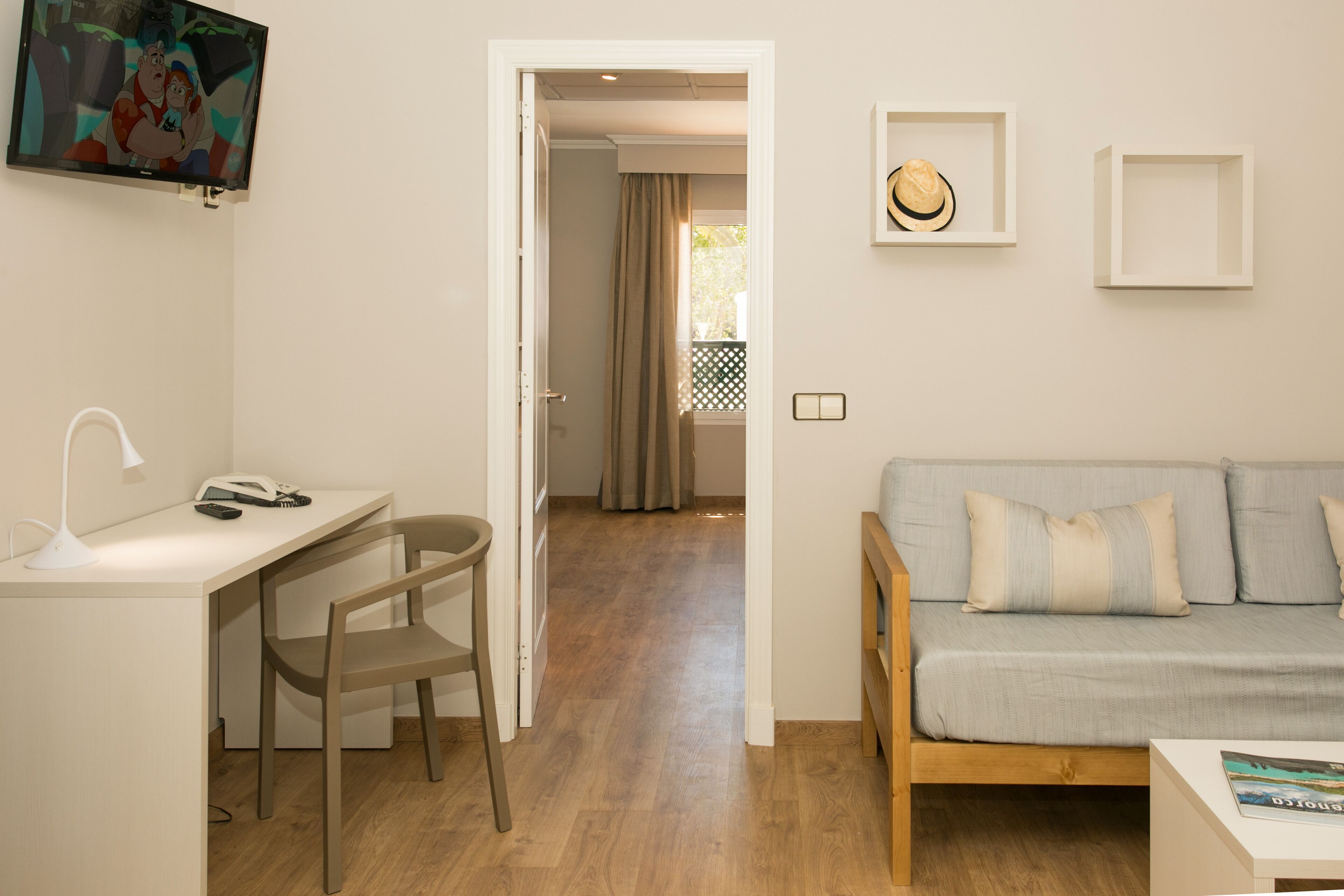 Una habitación con escritorio, silla y una fuente de luz. Hay un televisor montado en la pared, un sofá con cojines y una puerta que da a otra habitación.