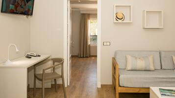 Una habitación con escritorio, silla y una fuente de luz. Hay un televisor montado en la pared, un sofá con cojines y una puerta que da a otra habitación.