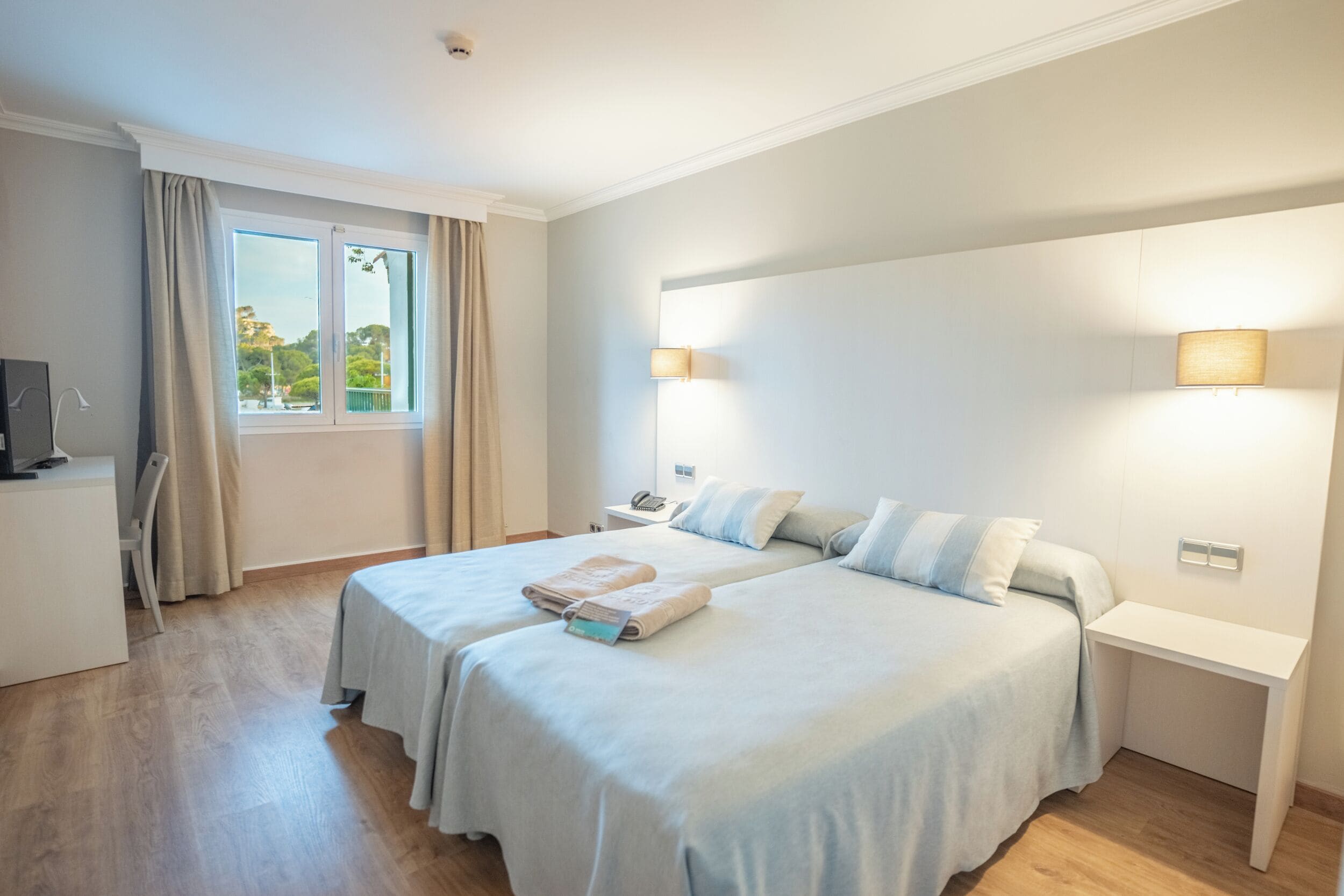A hotel room with two beds, a desk, a chair, a TV, and a window with curtains.