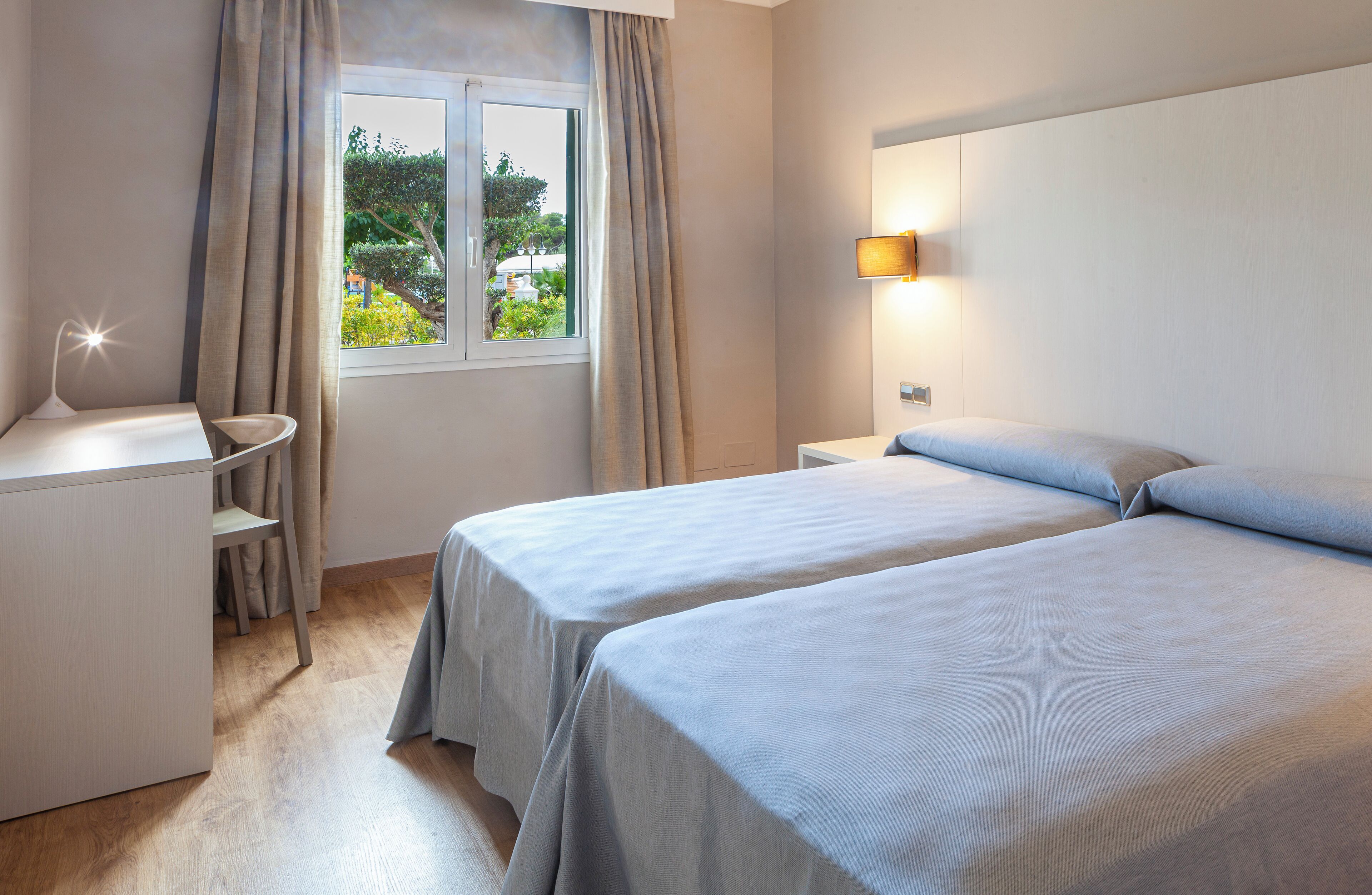 A hotel room with two beds, a desk with a lamp, a window with curtains, and a wall-mounted light.